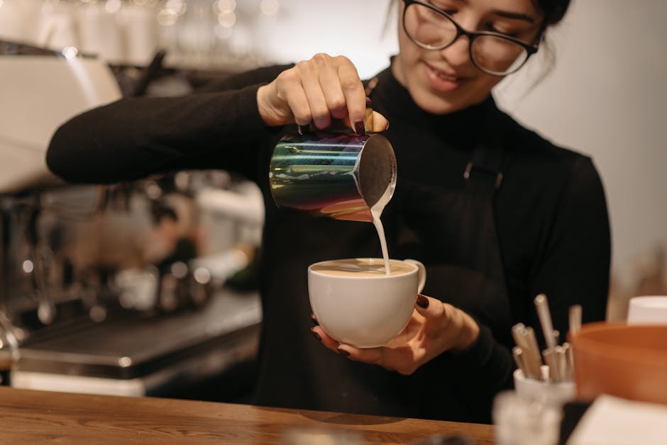 Barista steaming milk behind a coffee shop counter with customers in the background Barista steaming milk behind a coffee shop counter with customers in the background